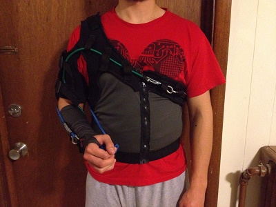 Front view of the sling including vest component, shoulder strap, arm sleeve, bands, belt loop holes, front zipper, forward facing adjustable arm straps, attachment point, and elastic band at bottom of torso portion. Tension distributed along neoprene (black) while athletic material (grey) is cooler and more comfortable.